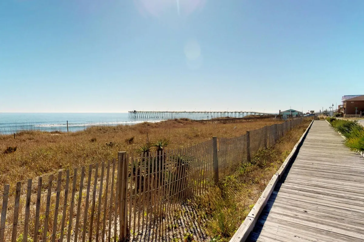 A tranquil view featuring the iconic Kure Beach pier stretching over calm ocean waters, with a sandy shoreline, grassy dunes, and a wooden boardwalk. The soft lighting suggests either sunrise or early morning, accentuating a serene and romantic beach atmosphere.