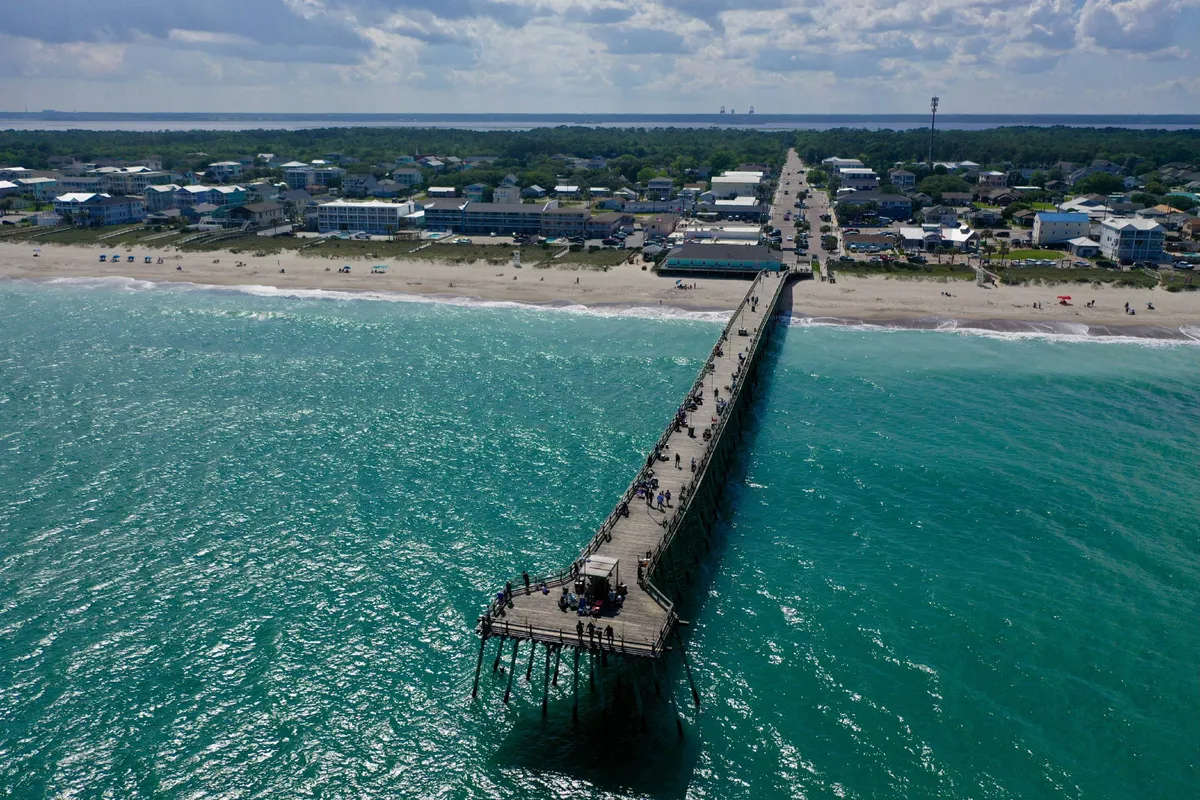 This image shows a lively beachside scene with a pier stretching into the ocean, sandy shoreline, and the nearby town visible under a partly cloudy sky. The presence of the pier, vast sandy beach, and open water captures the essence of coastal exploration and daily walks, which are ideal for pet-friendly adventures.