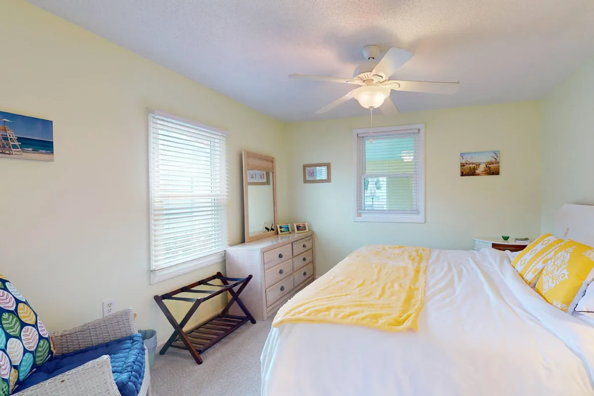 A peaceful bedroom features a bed with a yellow blanket, dresser, window with shaded blinds, and decorative photos on the walls. The soft morning light creates a tranquil, private retreat.