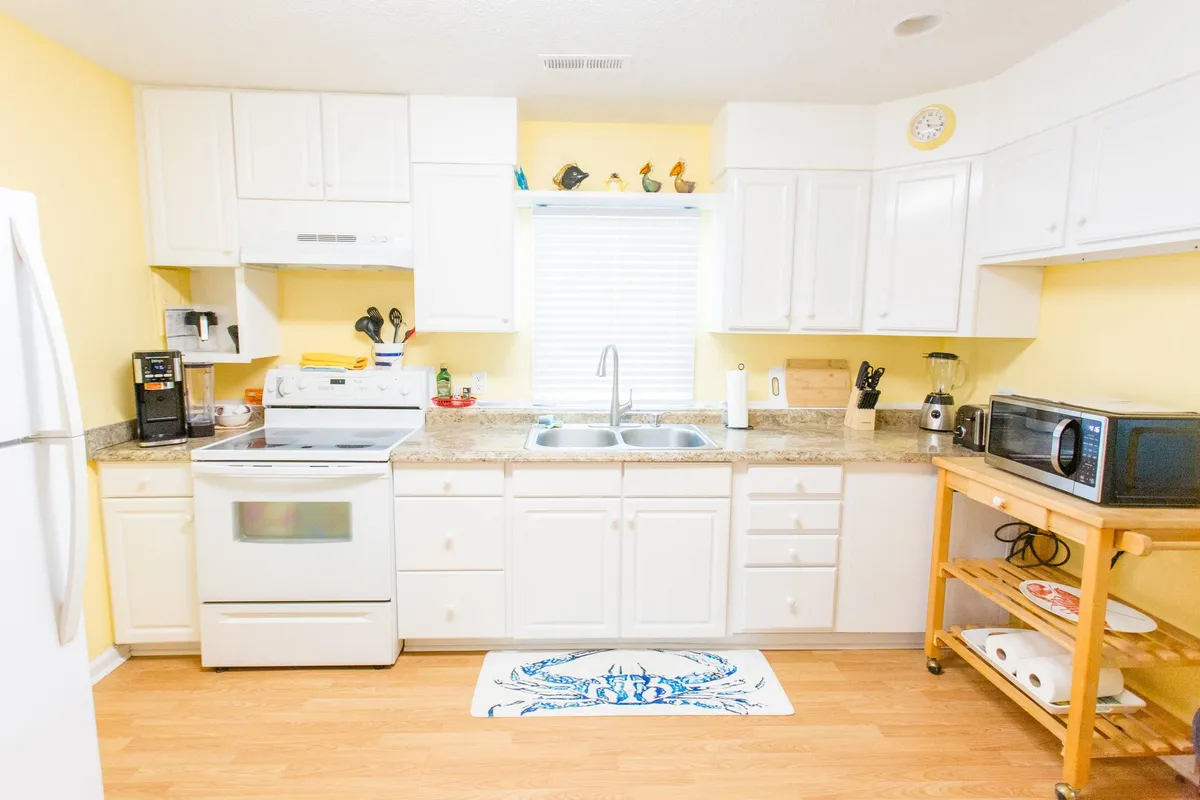 A bright kitchen with white cabinetry, countertop appliances, and a window that lets in cheerful morning sunlight. The kitchen appears well-equipped and inviting, with hints of fresh coffee and meal prep activities.