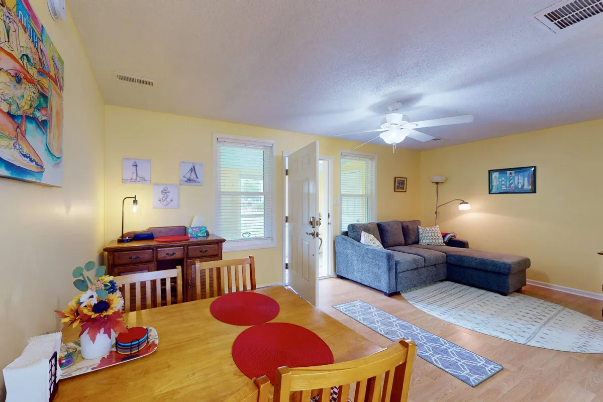 An open-plan space with a dining table set for four, a comfortable sofa, artwork on the wall, and the entrance door in view. The layout showcases the ease of movement and the seamless connection between dining and living areas.