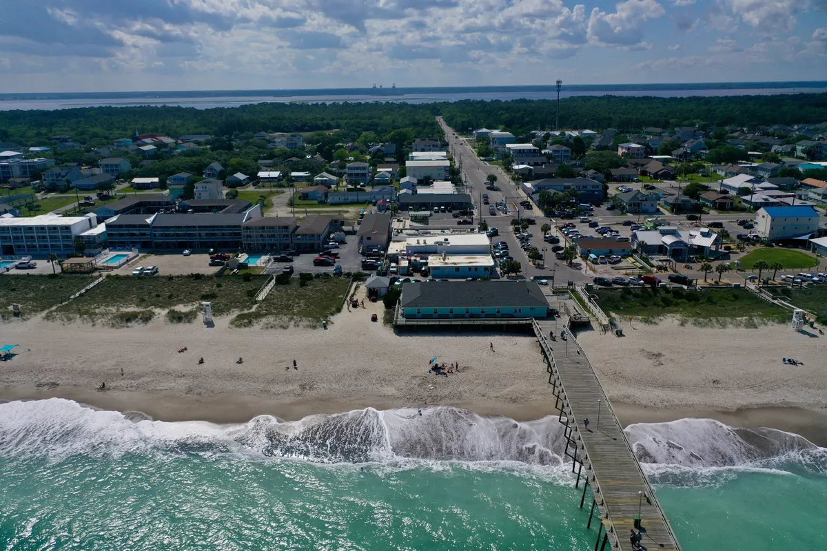 A panoramic view of the beach with the pier extending into the ocean, nearby town buildings, and a lively yet relaxed atmosphere. The scene shows multiple activities including walking, fishing, and exploring the local shops and dining spots that are visible along the shore.
