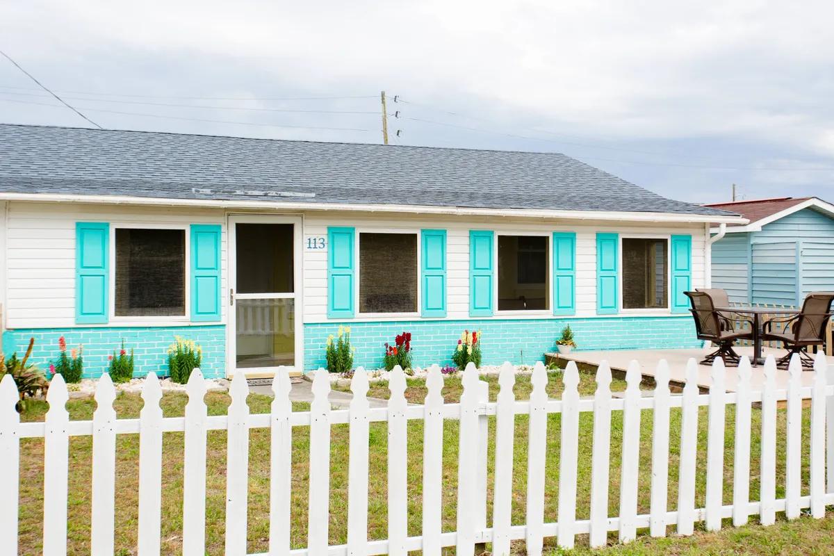 An inviting exterior view of Bec's Beach Bungalow, with cheerful turquoise shutters, a classic white picket fence, and blooming flower beds in the foreground. The private yard is visible, suggesting a safe space for children to play and families to relax after a day out.