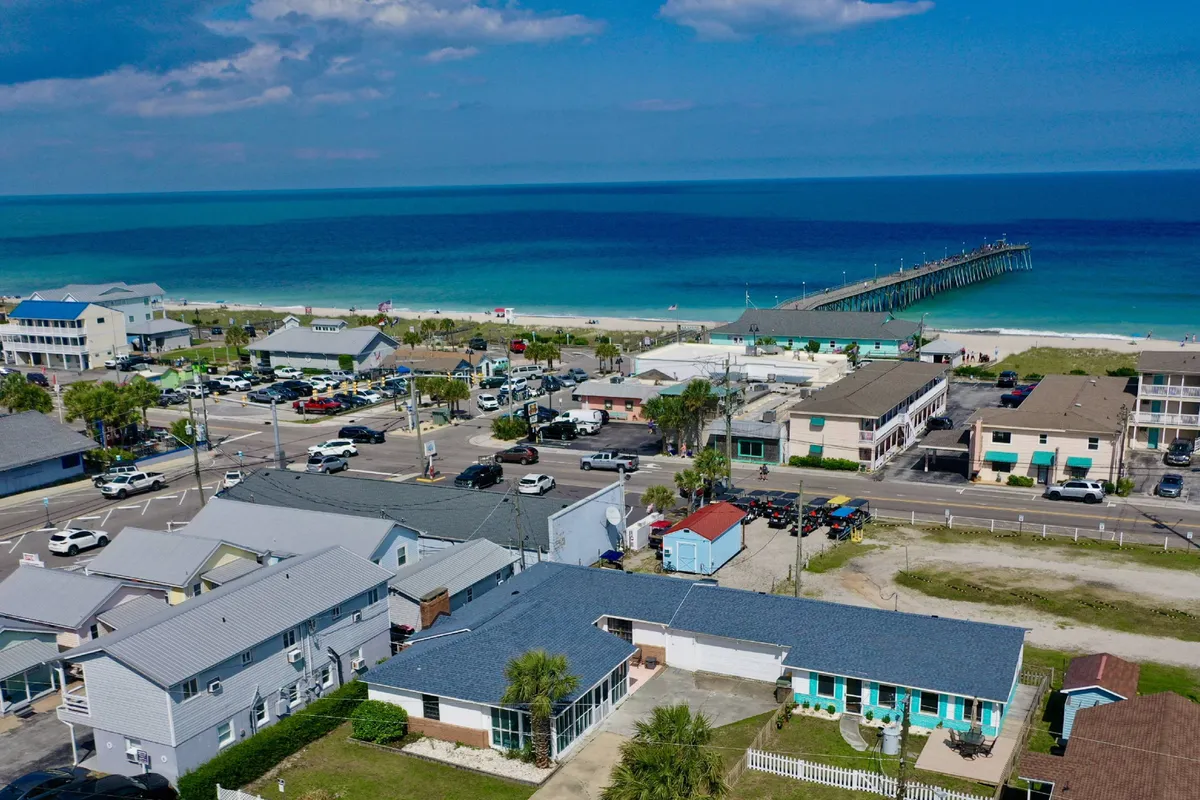 Overlooking the coastline, this image features the beach, a pier, ocean waves, residential buildings, and a parking area, suggesting easy access for outings. The scene is casual and welcoming, inviting visitors to enjoy beach activities with their dogs.