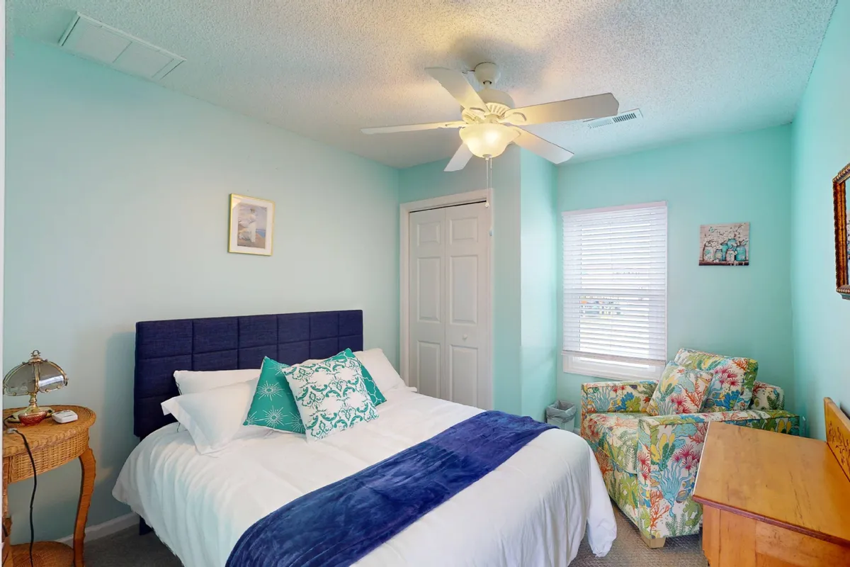 A calm and serene bedroom with a bed adorned with blue and green pillows, a colorful armchair, a wooden dresser, and framed artwork. Natural light filters into the space, emphasizing its welcoming environment.