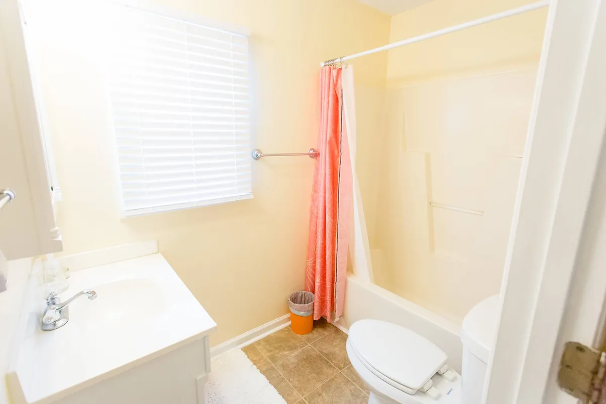 A bright, inviting bathroom with a sink, shower area, toilet, and a window with blinds. The room feels fresh and clean, perfect for starting or ending the day comfortably.