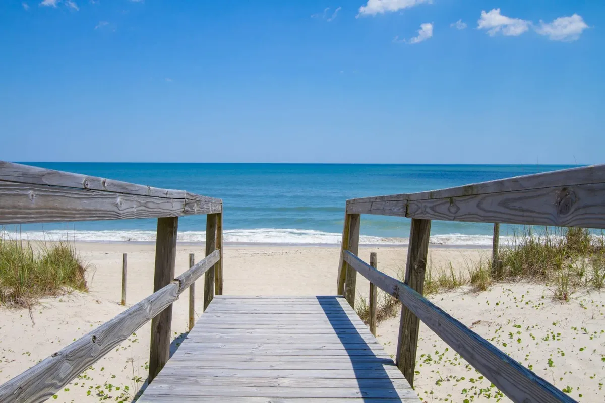 This image highlights a wooden walkway leading directly to the sandy beach and ocean beyond, with a clear sky above. The path suggests inviting, direct access for guests and their pets, with space for off-leash play and scenic walks.
