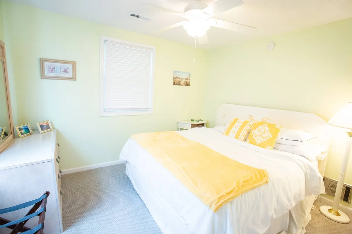 A tranquil bedroom featuring a bed with a yellow blanket and pillows, a dresser with a mirror, and a lamp softly illuminating the space. The arrangement creates a peaceful atmosphere ideal for rest and relaxation.