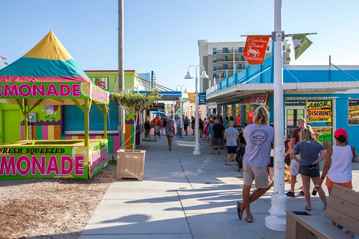 A vibrant scene depicting a bustling boardwalk area with a colorful lemonade stand, crowds of people, and bright shops, encapsulating the festive, communal spirit of beach vacations.
