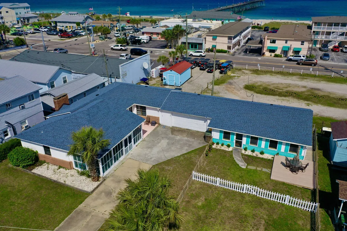 A relaxed patio area with a blue roof overlooks a green lawn, featuring a comfortable space for outdoor dining and barbecues. The view also hints at proximity to the beach and pier.