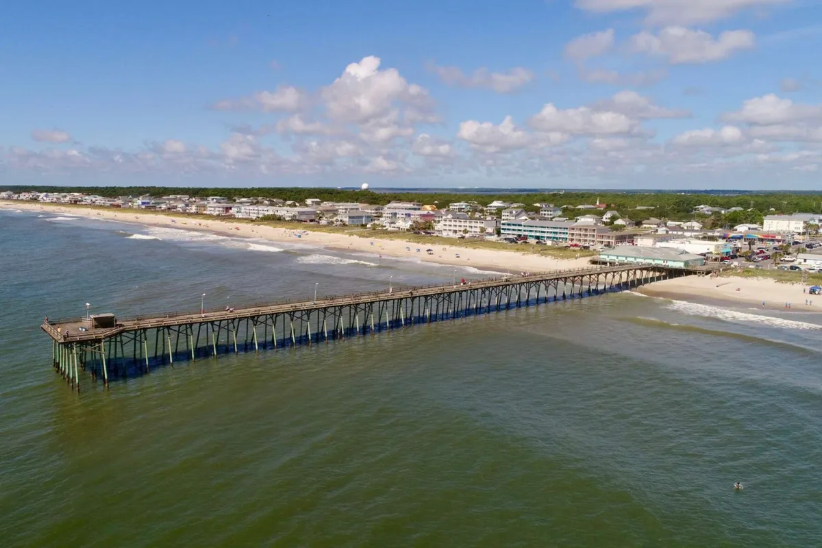 A sweeping view of the Kure Beach pier stretching over the ocean, bordered by a wide sandy beach where children and families can be seen playing, with charming coastal houses and soft clouds in the background. The scene evokes a family-friendly shoreline ideal for sandcastle building and splashing in the waves.