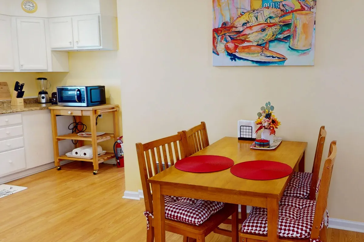 A welcoming interior dining area with a wooden table surrounded by chairs, colorful artwork on the wall, and visible kitchen appliances in the background. The space is set up for meals and gatherings, with warm lighting creating a cozy atmosphere.