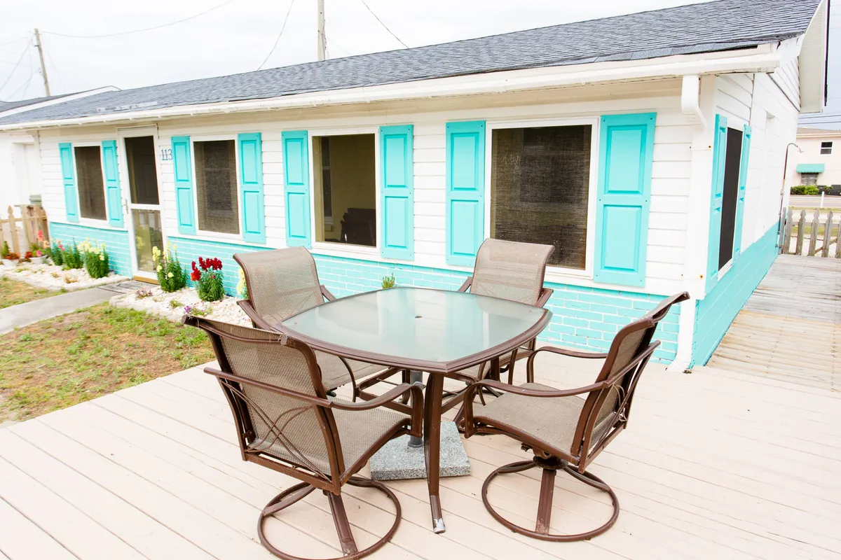 This image captures a cheerful outdoor dining table set on a sunny deck, surrounded by brightly colored shutters and a well-kept landscaped garden. The scene is inviting, perfect for al fresco meals and casual gatherings.