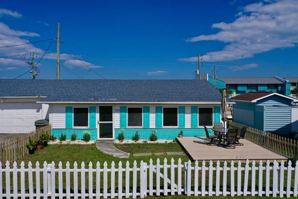 The image features a charming beach bungalow with cheerful turquoise shutters, a classic white picket fence, and a welcoming deck adorned with comfortable seating. The setting is bright and inviting, capturing a golden sunset that hints at relaxing evenings outdoors. The front garden is tidy and lush, completing the picture-perfect coastal vibe.